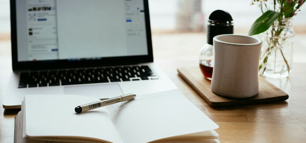 a classic content creation desk set up with a notebook, pen, laptop, plant and coffee mug